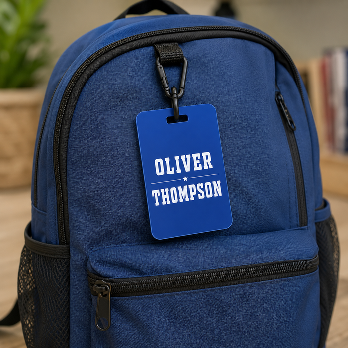 A blue canvas backpack with black zippers and side mesh pockets, featuring a personalized blue luggage tag that reads 'OLIVER THOMPSON' attached near the top zipper.