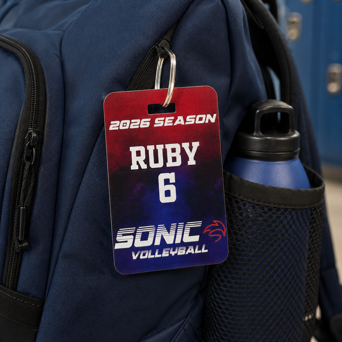 A close-up of a navy blue backpack with a red-to-purple gradient season pass tag attached near the zipper. The tag reads '2026 SEASON', 'RUBY', '6', and 'SONIC VOLLEYBALL'.