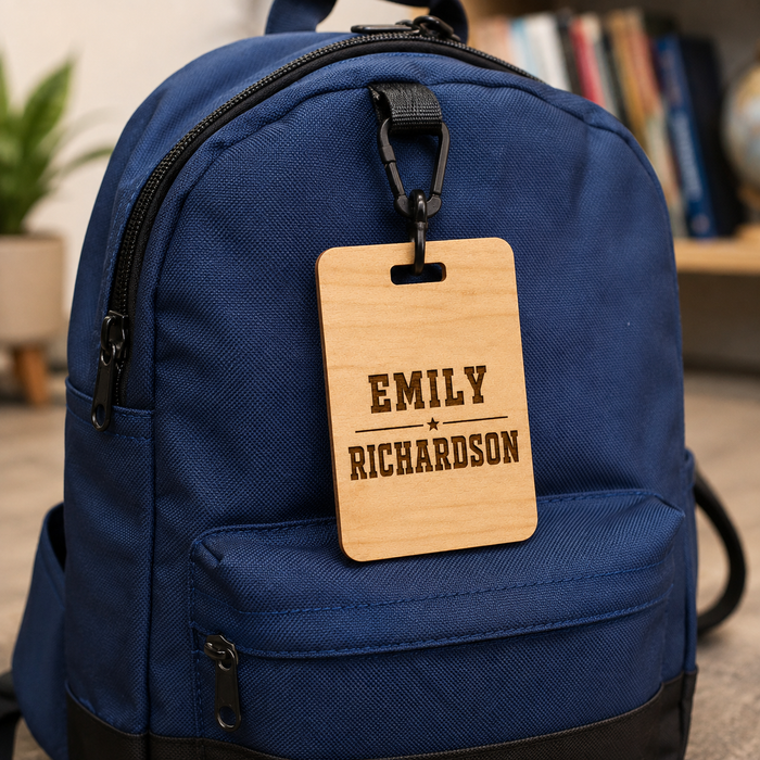 A navy blue backpack with a personalized wooden luggage tag hanging from a black carabiner. The tag is engraved with the names 'EMILY' and 'RICHARDSON' separated by a star.