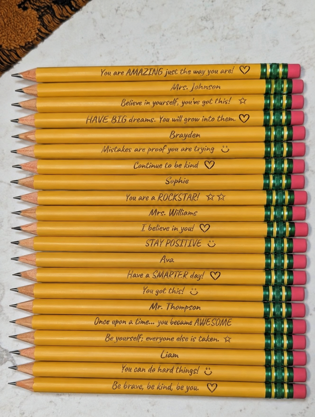 A stack of personalized yellow pencils with handwritten motivational quotes and heart icons, arranged vertically on a light marble surface.