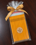 A gift set of 12 personalized yellow pencils wrapped in clear cellophane and tied with a gold ribbon, displayed on a dark wood background.
