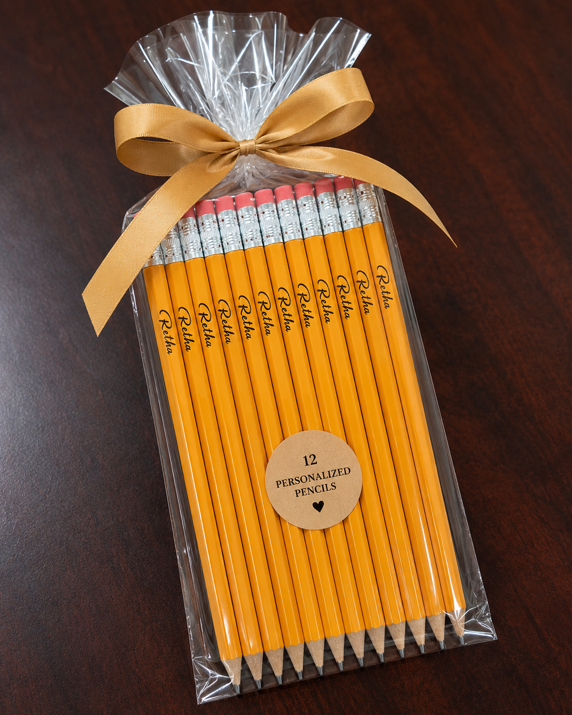 A gift set of 12 personalized yellow pencils wrapped in clear cellophane and tied with a gold ribbon, displayed on a dark wood background.