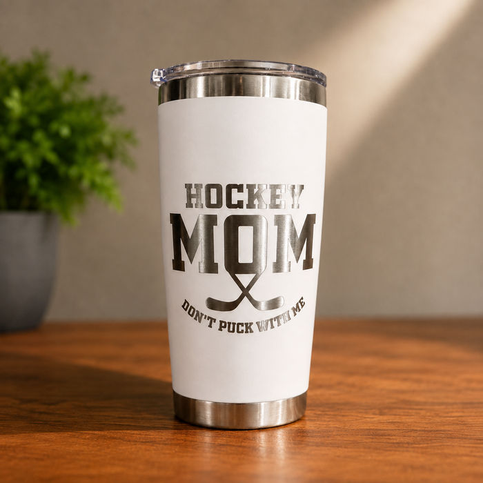 A white insulated travel tumbler with a clear lid sits on a wooden table. The tumbler features a black graphic print that reads 'HOCKEY MOM' with crossed hockey sticks and 'DON'T PUCK WITH ME' underneath.